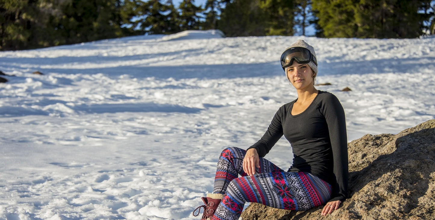 Person sitting on a rock in a snowy landscape wearing ski goggles and patterned pants.