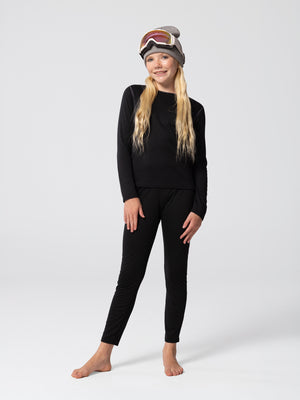 Full-body view of youth wearing Hot Chillys Pepper Bi-Ply black thermal crewneck top and matching bottoms, with a beanie and goggles