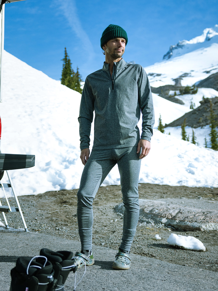 Man standing outdoors on a snowy mountain wearing Hot Chillys Micro-Elite Chamois heather gray zip-neck top and tights with a green beanie