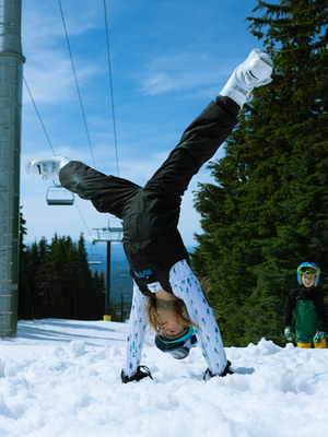 Youth doing a handstand in the snow, wearing Hot Chillys Micro-Elite Chamois Downhill Ski print leggings visible under a black ski jacket, with ski goggles and boots