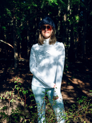 Woman standing in a sunlit forest wearing a white Hot Chillys Micro-Elite Chamois base layer top with light-colored leggings, a dark cap and sunglasses