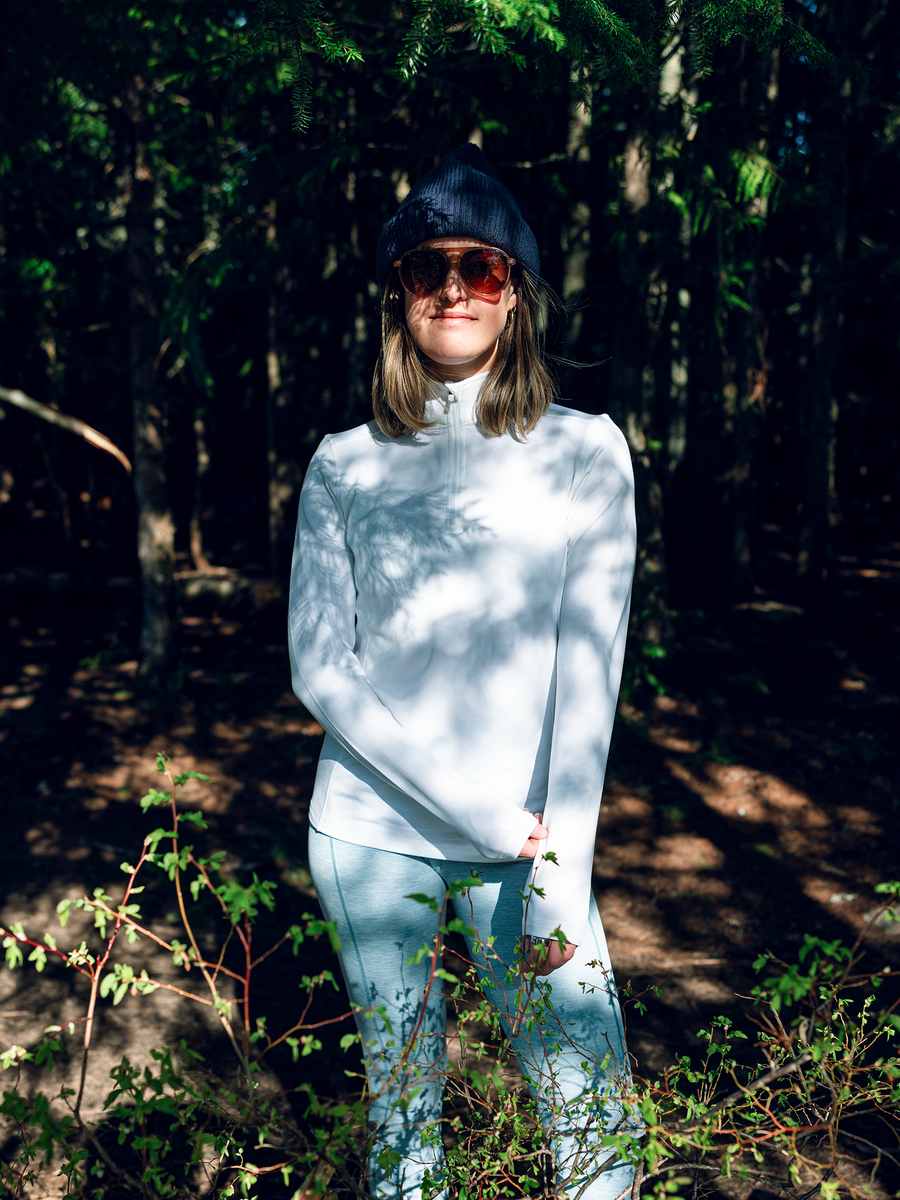 Woman standing in a sunlit forest wearing a white Hot Chillys Micro-Elite Chamois base layer top with light-colored leggings, a dark cap and sunglasses
