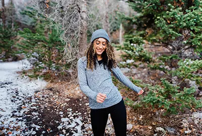 Woman in a gray knit hat and light blue sweater standing in a snowy forest.