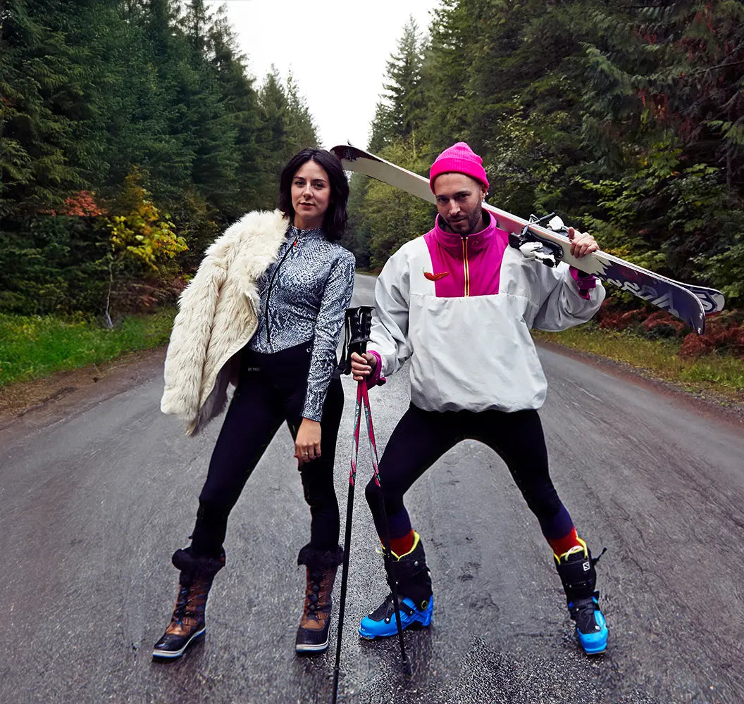 Two people with skis on a road surrounded by trees